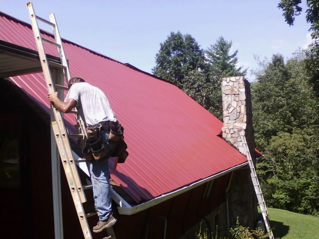 Roofer on a ladder beside a red metal roof and stone chimney — Soffit & Fascia Repair by Roofing Brothers in Fayette
